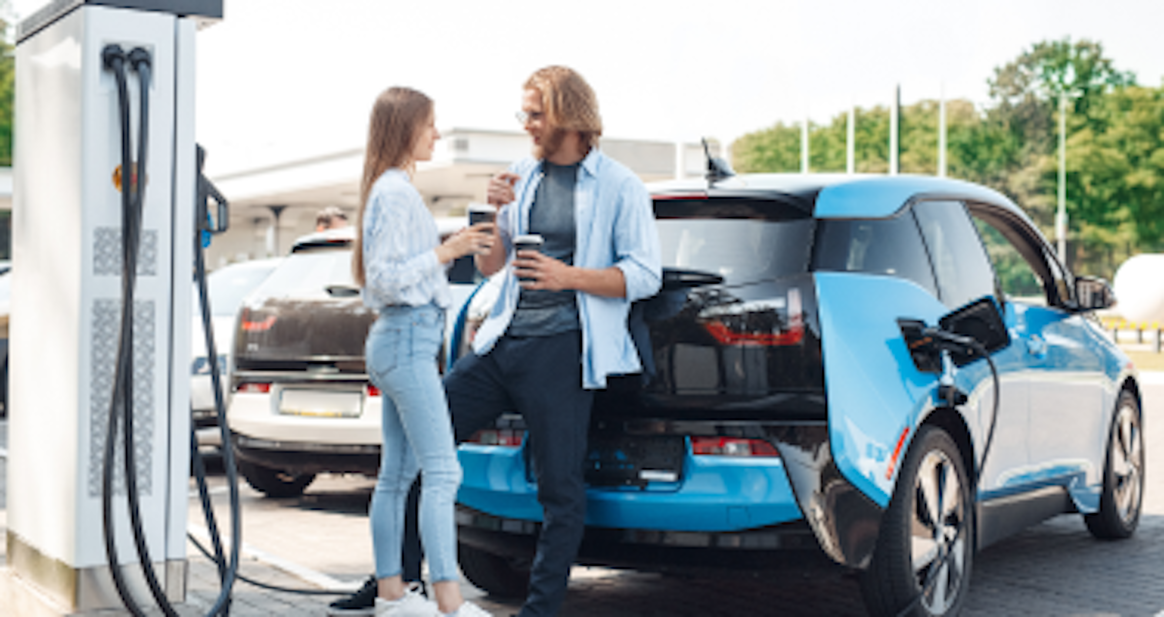 A man and woman chat by an electric car at a charging station.