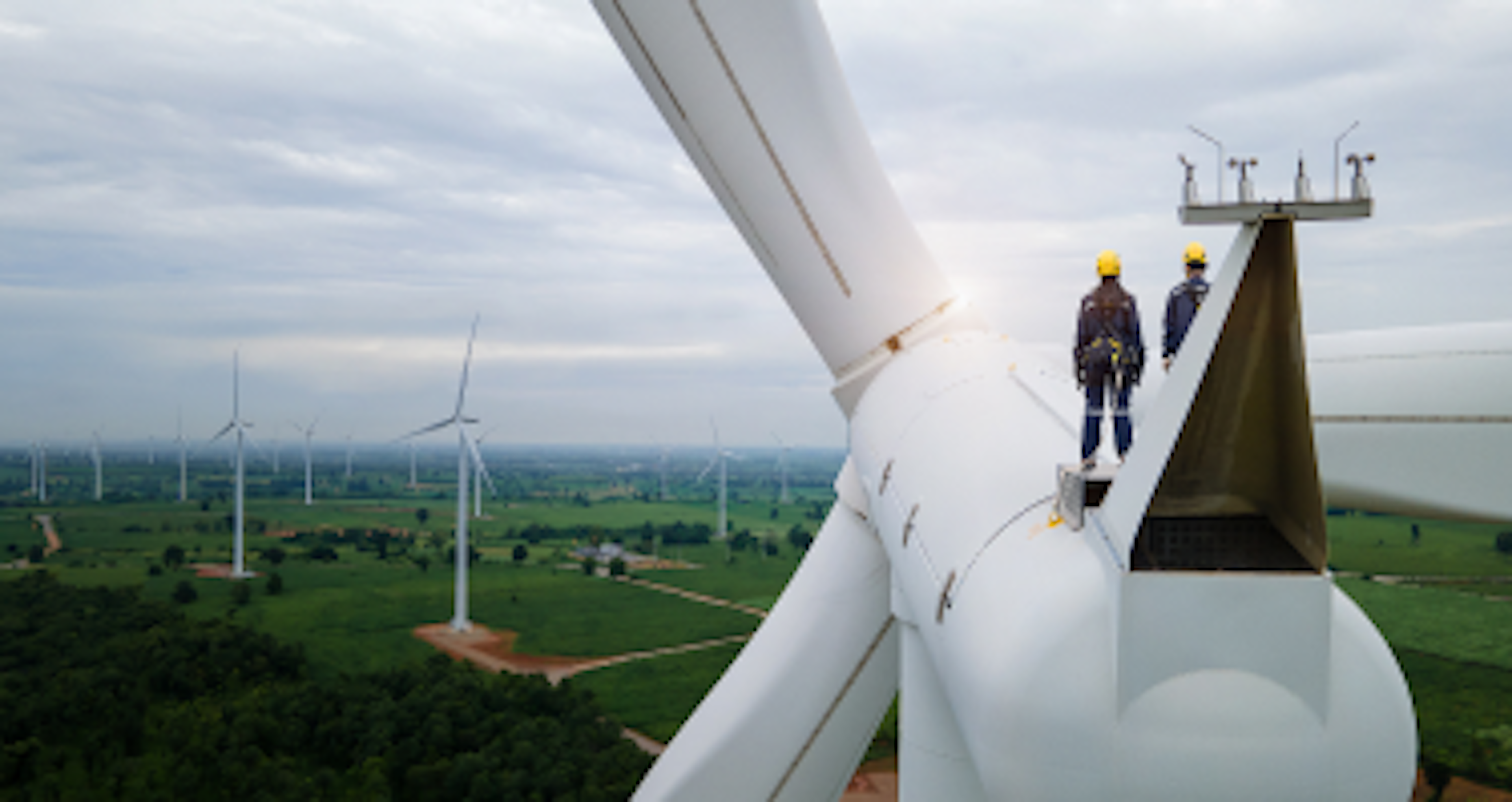 Two technicians in safety gear stand on a wind turbine, overlooking a wind farm under a cloudy sky.