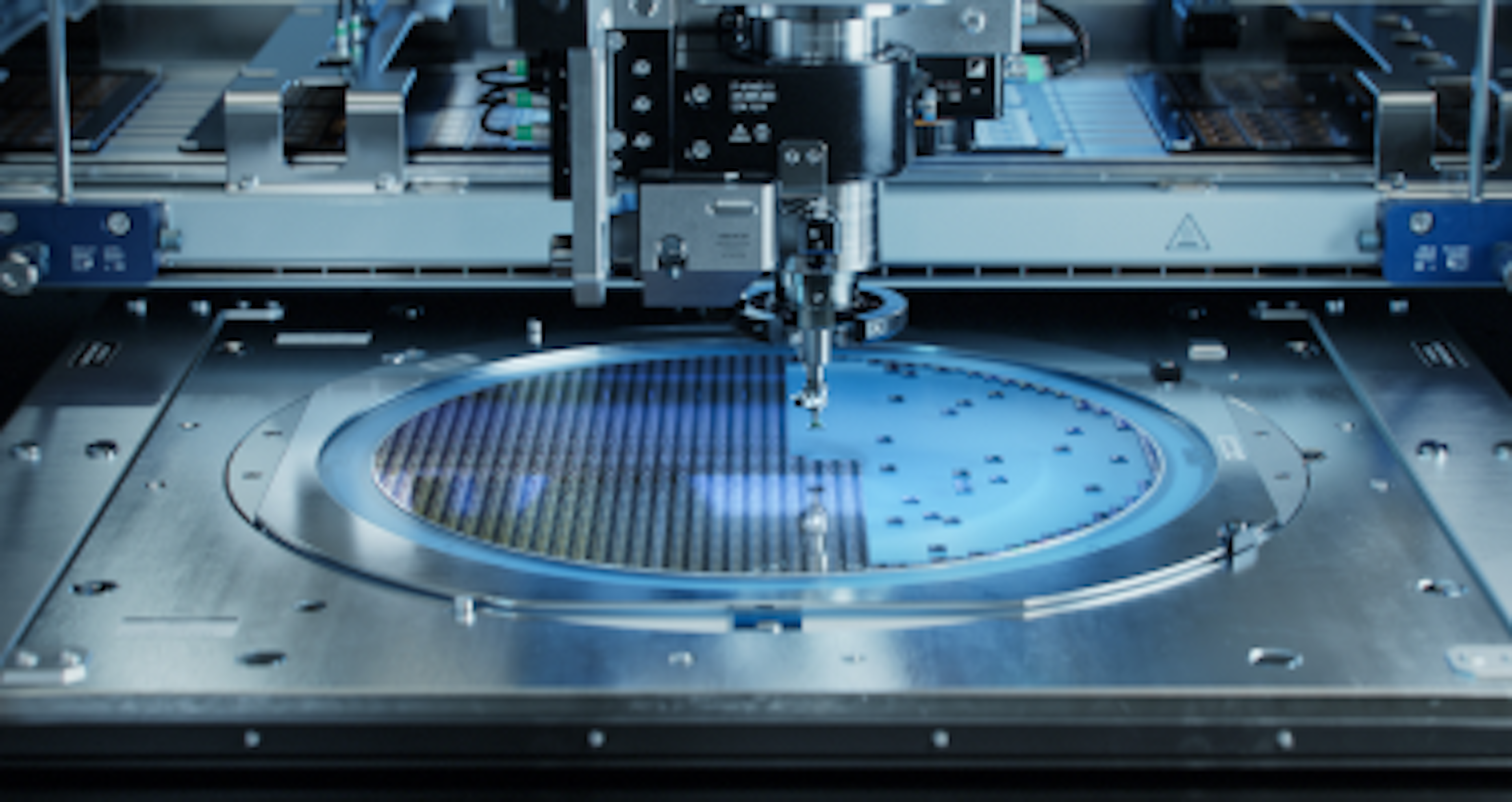 The image shows a silicon wafer being worked on by a precision machine in a cleanroom.