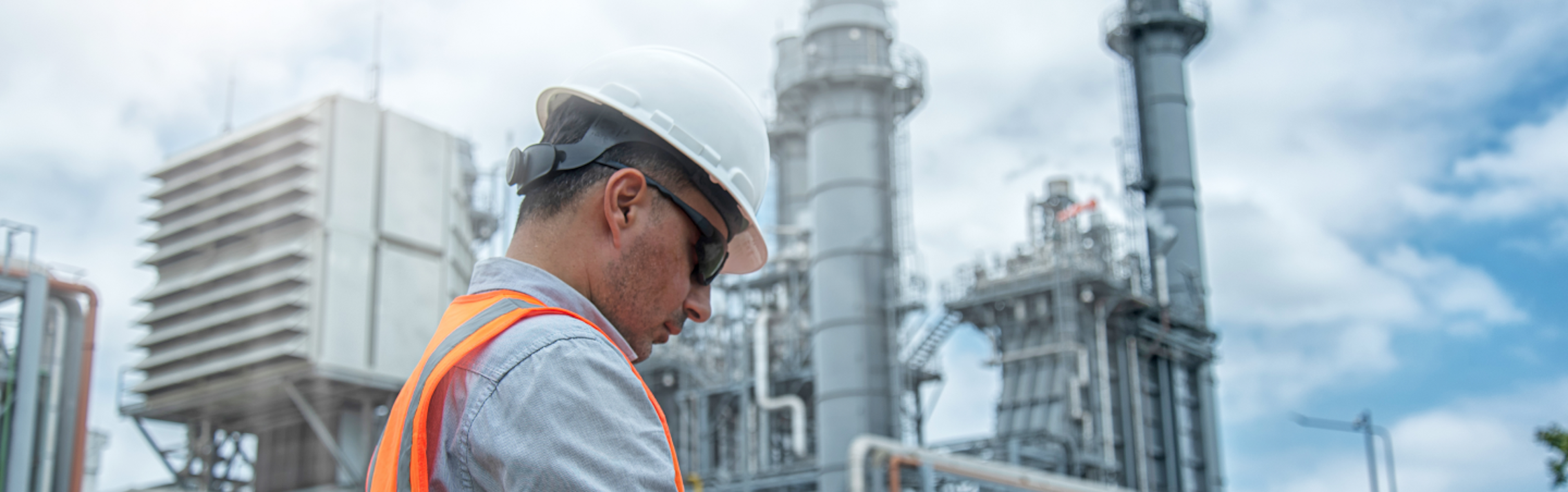 A person in a hard hat and vest facing industrial structures. The sky is partly cloudy, and their face is pixelated.