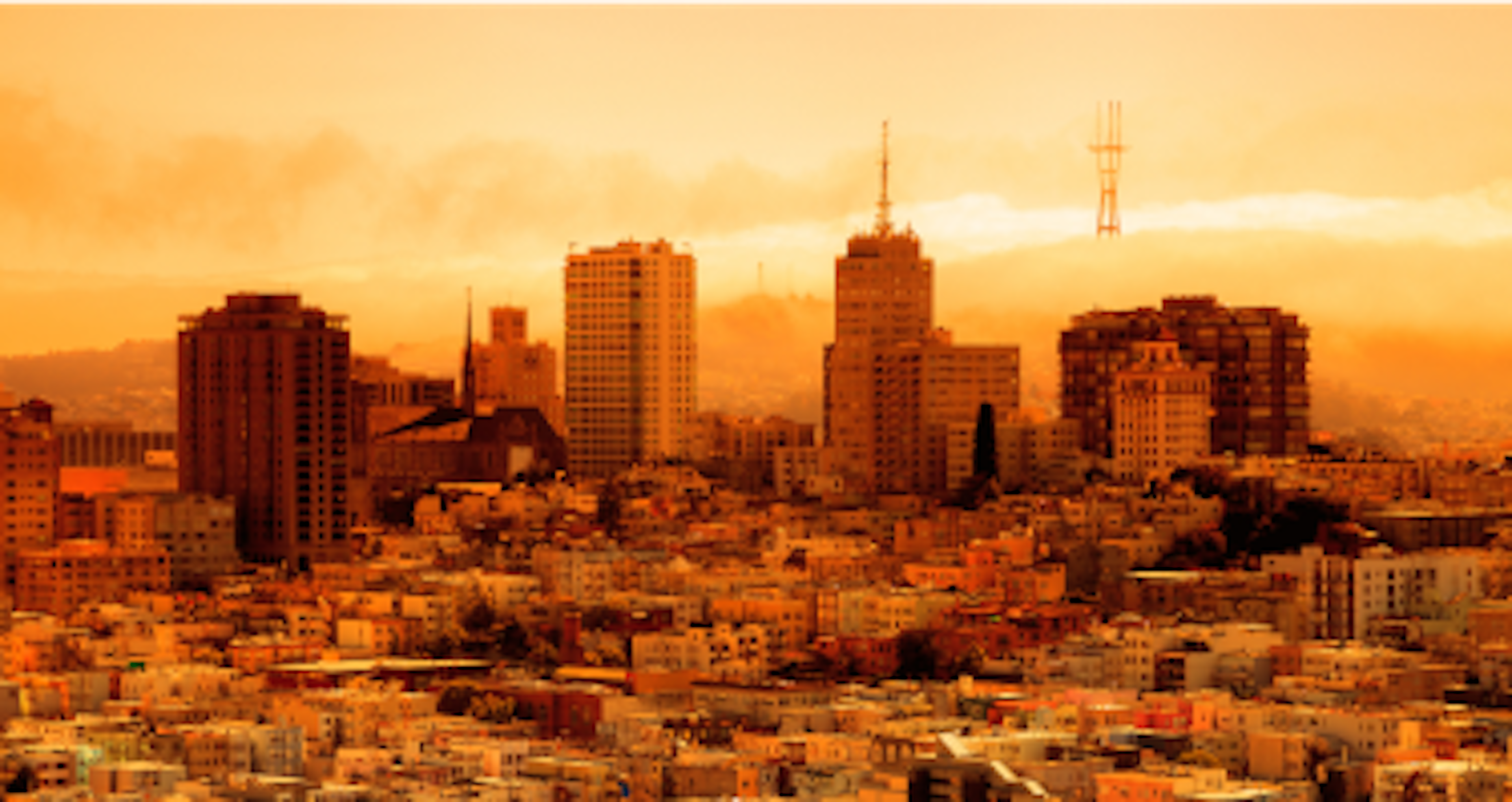 A city skyline at sunrise or sunset, with high-rise buildings and antenna towers against a hazy, orange sky.