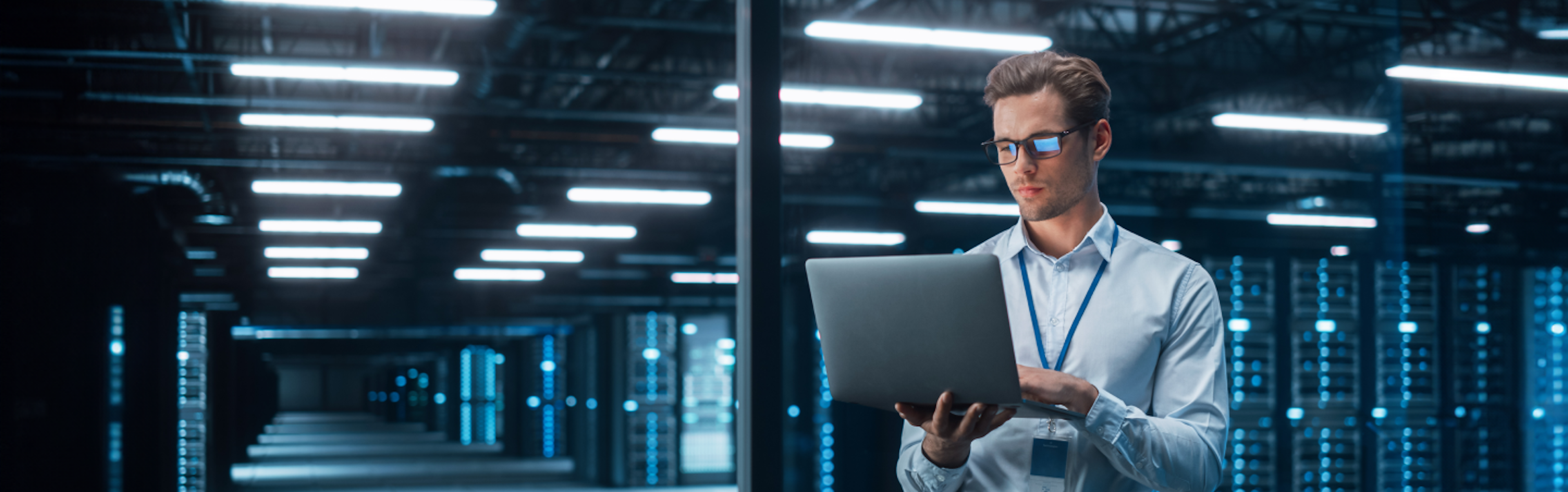 A man in a white shirt and glasses stands in a dimly lit server room, working on a laptop. He wears a lanyard and looks concentrated. Rows of server racks with blue lights are visible behind him.