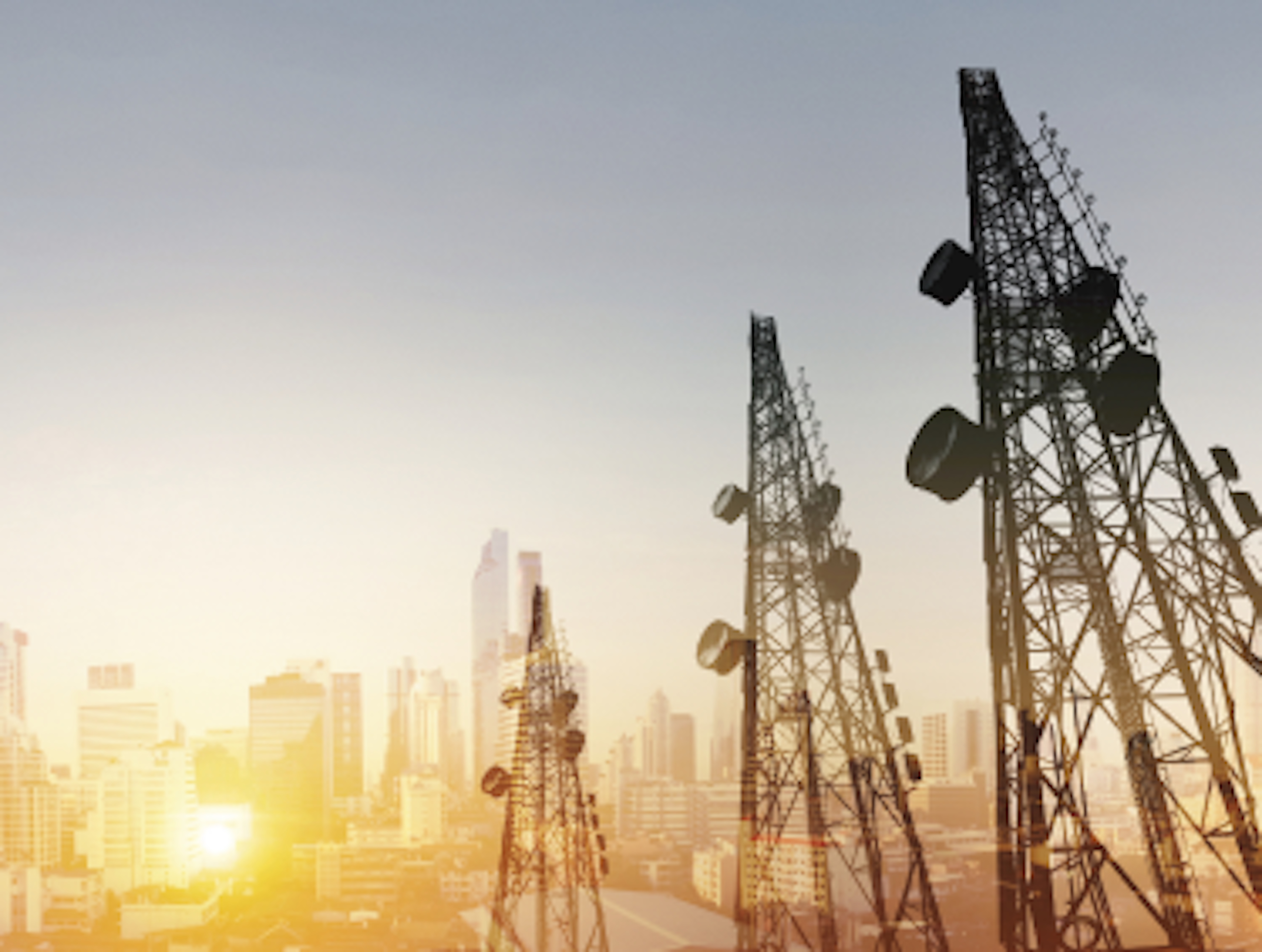 Skyline view of a city at sunset with multiple large telecommunication towers in the foreground.