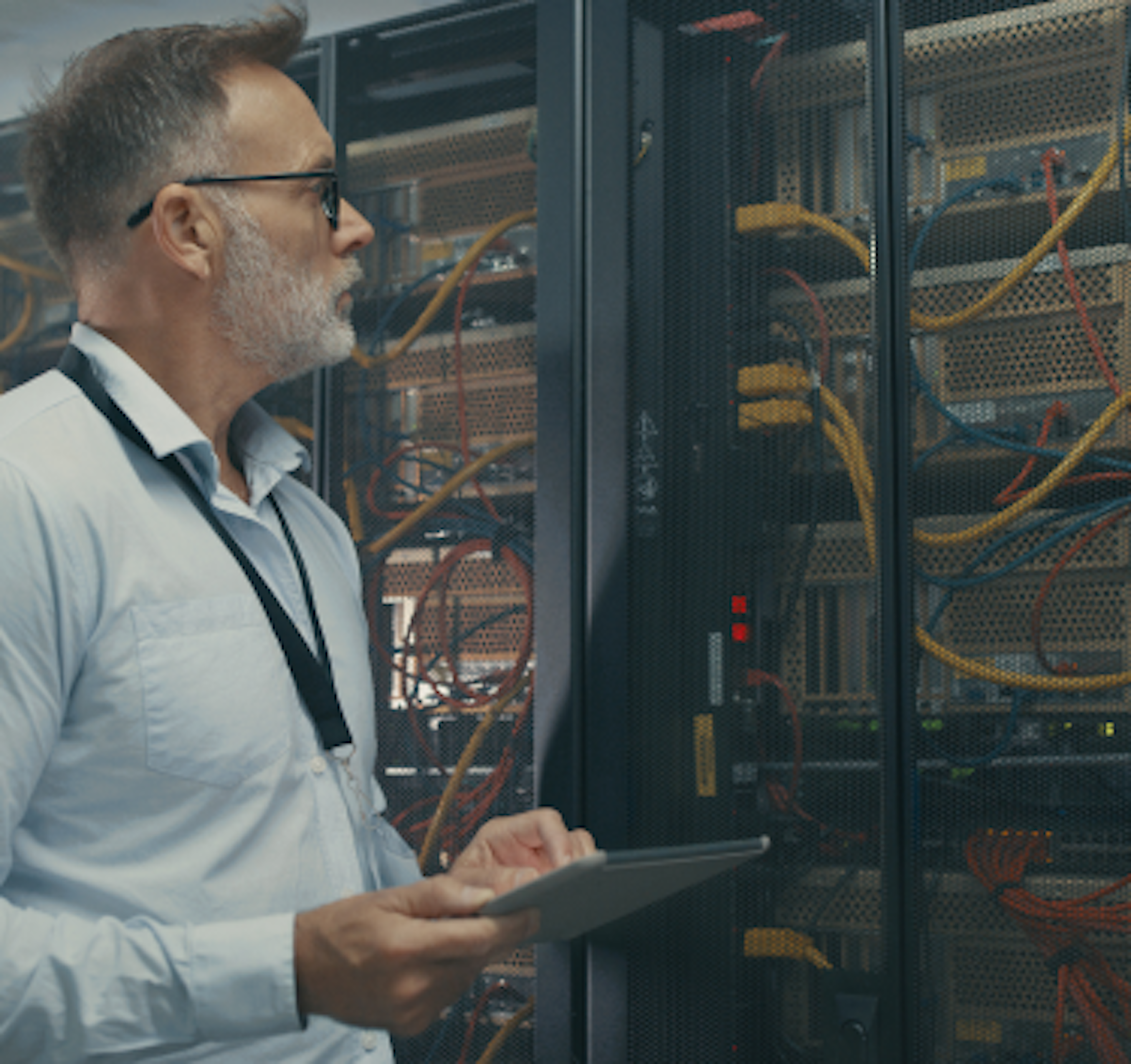 A man with glasses and a beard examines server racks in a data center, holding a tablet and wearing a lanyard, surrounded by networking cables and equipment.