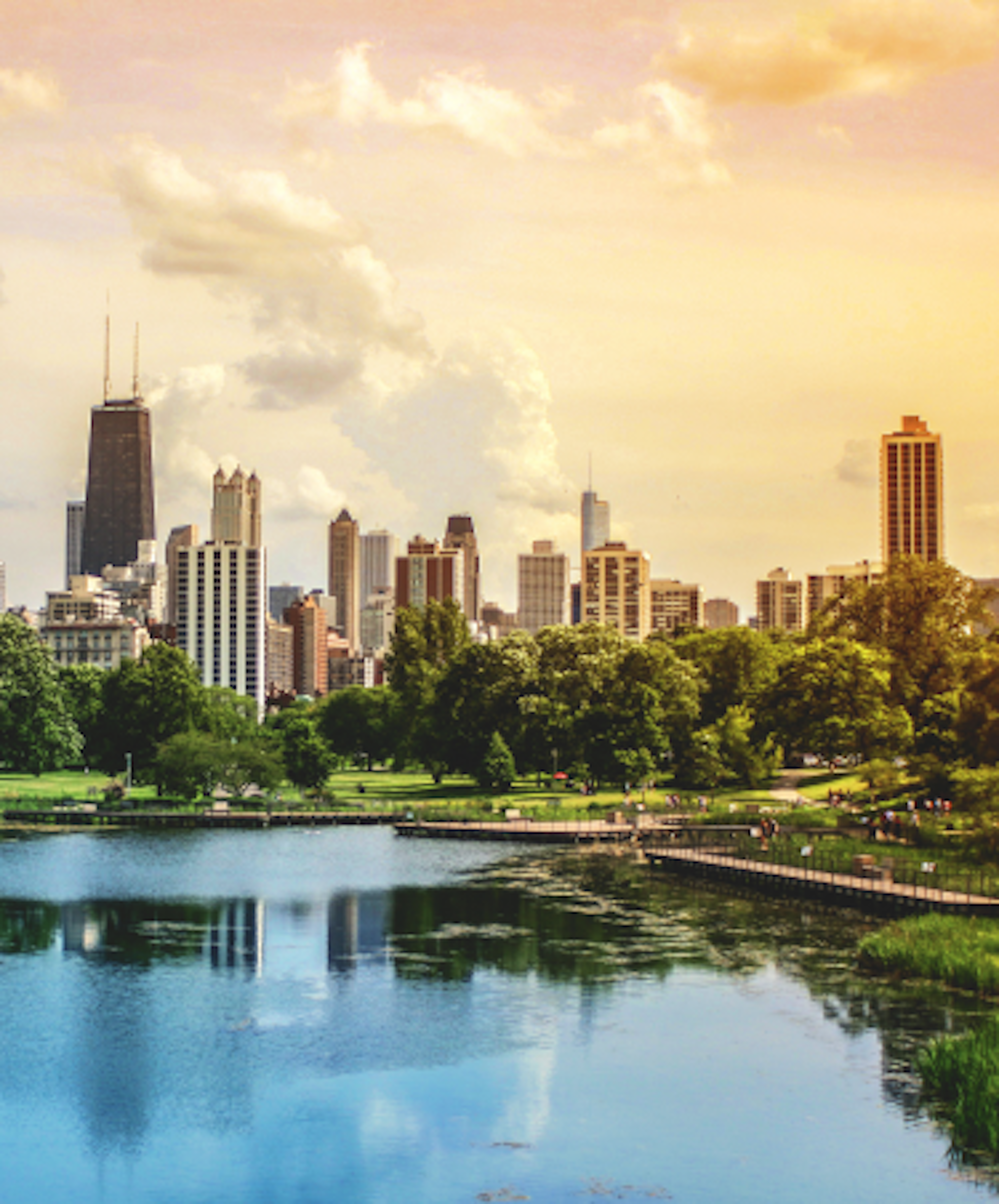 A city skyline with tall buildings is seen from across a green park under a cloudy sky.