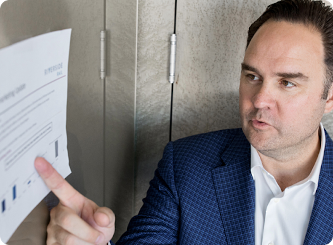 A man in a suit points at a document with graphs and text attached to a wall.