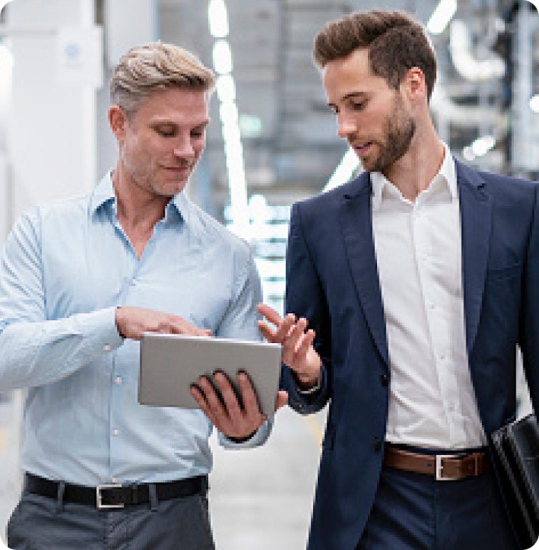 Two men in formal attire discuss something on a tablet while walking in a well-lit industrial or office space.