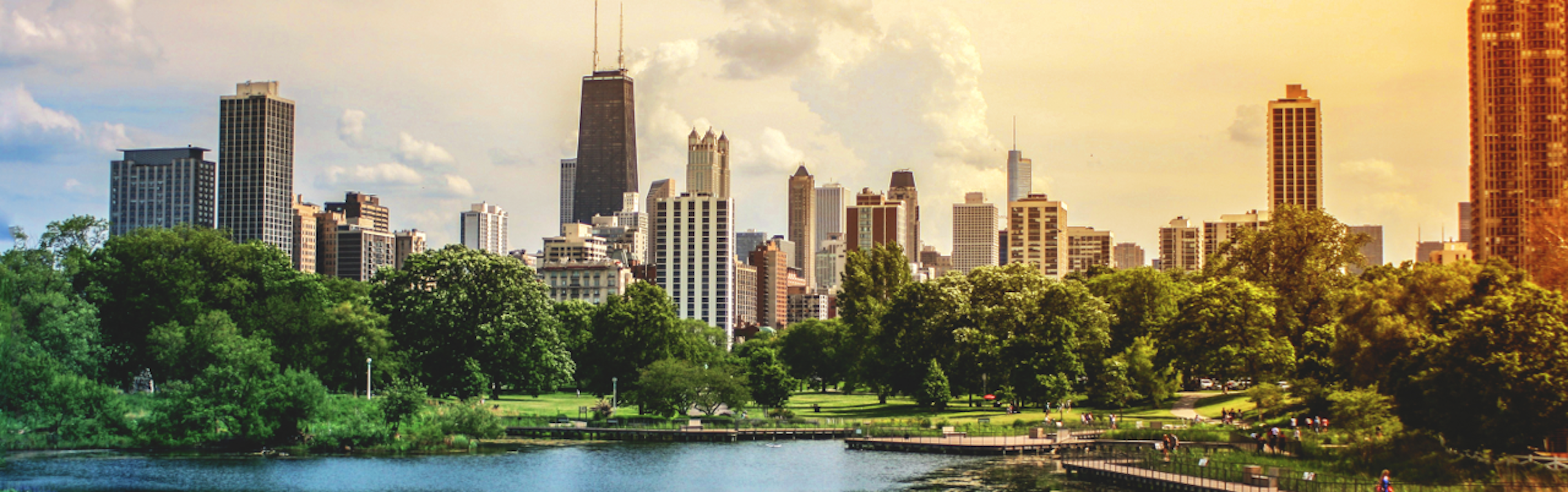 A city skyline with tall buildings is seen from across a green park under a cloudy sky.