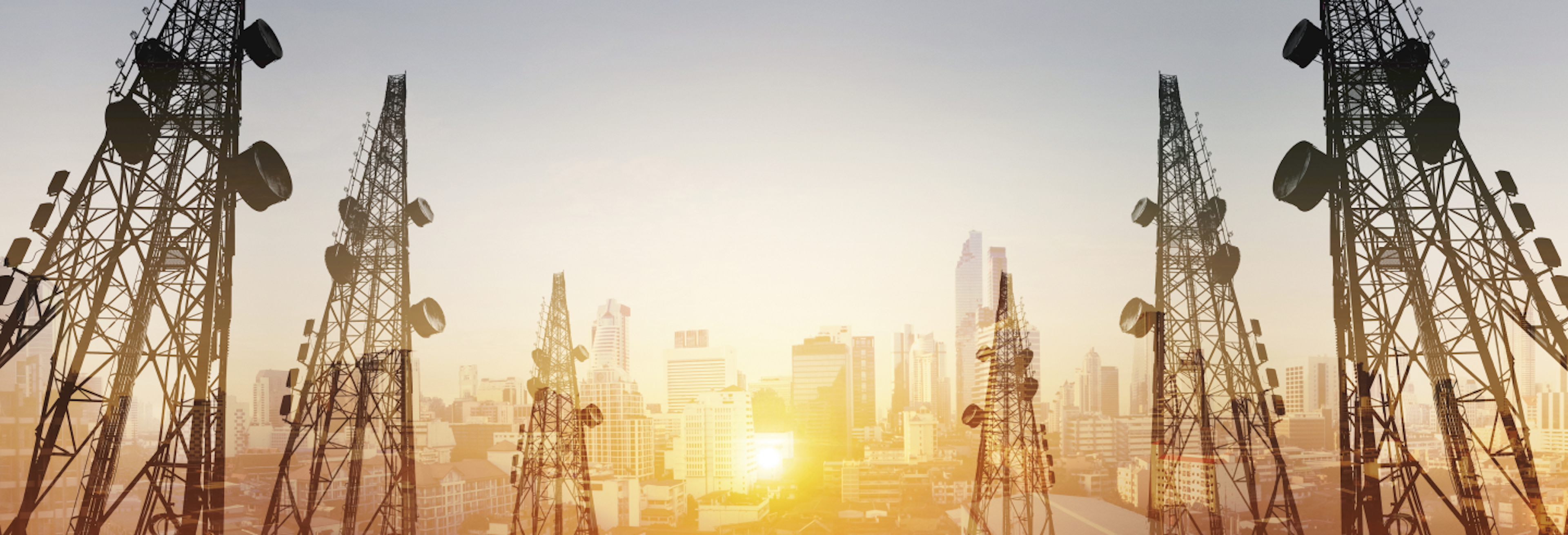 Skyline view of a city at sunset with multiple large telecommunication towers in the foreground.